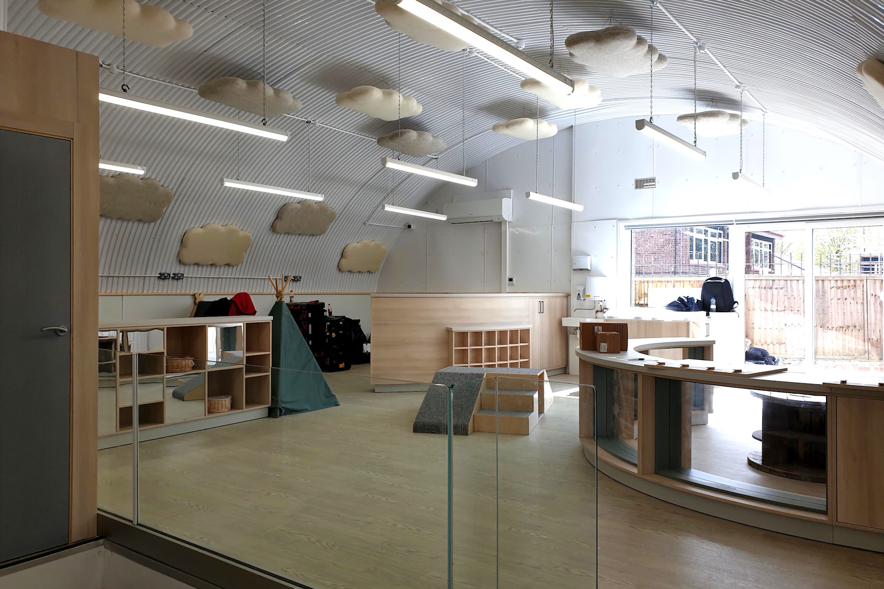 A modern classroom with curved ceiling, light wood furniture, play structures, shelves, and large windows letting in natural light.