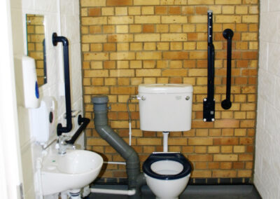 Accessible toilet with a loo, small basin, exposed plumbing, brick wall, and multiple black grab rails for support.