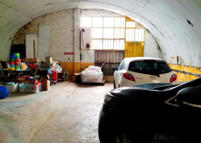Two cars are parked inside a curved-roof garage with yellow and white walls; various boxes and items are stored along the left side.