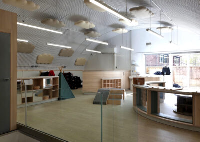 A modern classroom with curved ceiling, cloud-shaped decorations, wooden furniture, play equipment, and large windows letting in natural light.