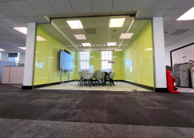 A modern glass-walled meeting room with a round table and chairs, a digital screen, and yellow-green walls, in a spacious office setting.