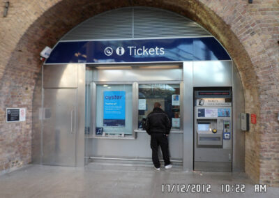 A person stands at a railway station ticket counter under a blue "Tickets" sign, with a ticket machine and informational posters nearby.