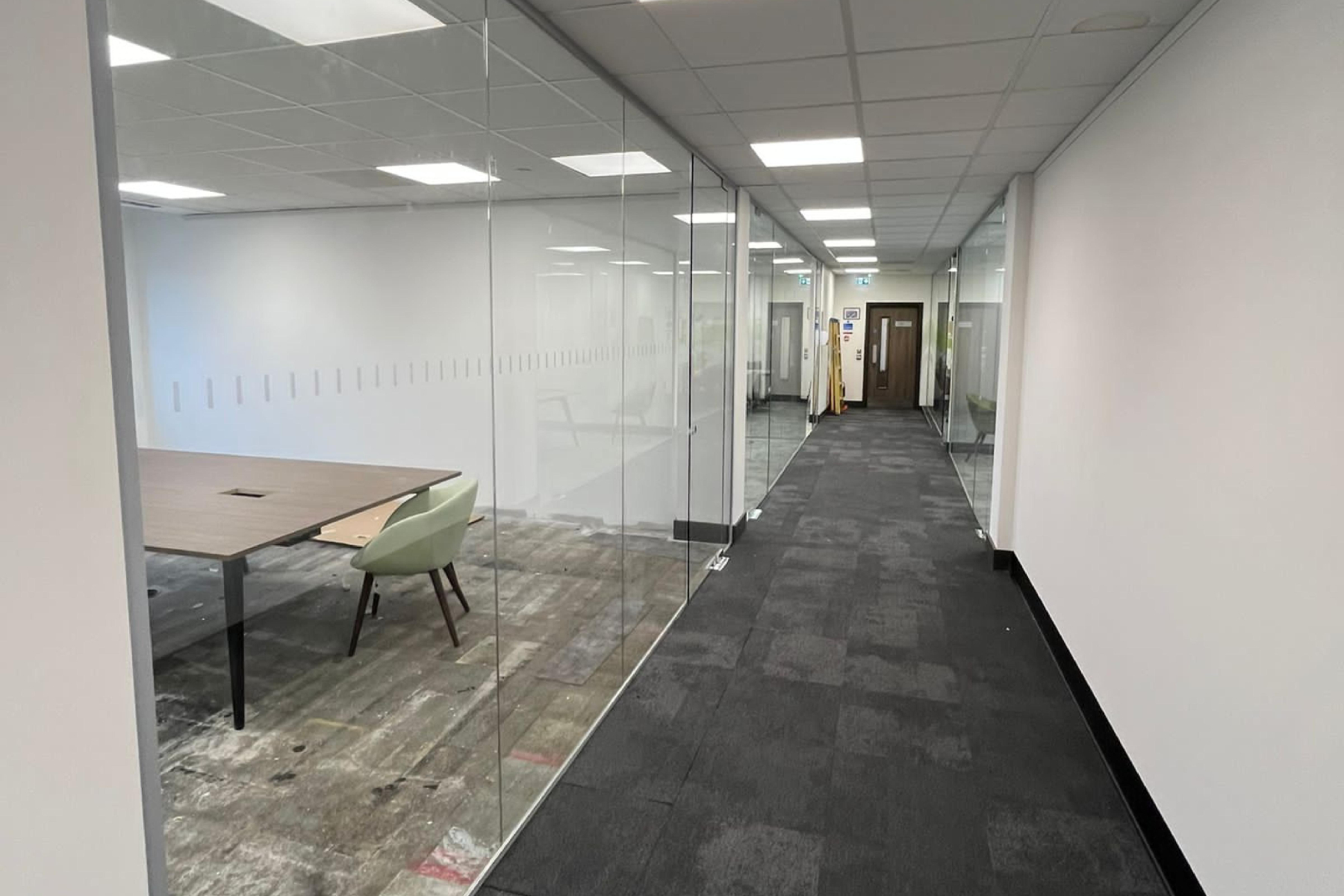 A modern office corridor with glass walls on one side, showing an empty meeting room with a table and chairs, and closed wooden doors at the end of the corridor.