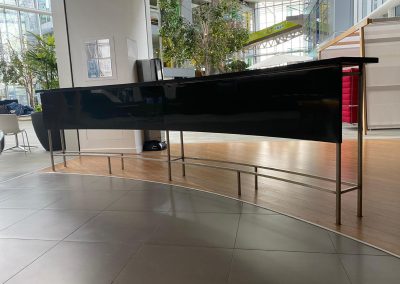 A long, black, glossy counter with metal legs stands on a curved floor transition between tiled and wooden surfaces in a modern indoor space with large windows and plants.