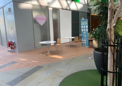 Two round tables with chairs are spaced apart in front of a partition labelled "PHYSICAL." A health and safety poster and fire extinguishers are visible nearby inside a modern building.