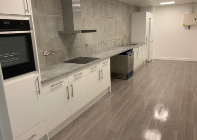 Modern kitchen with white cupboards, built-in oven, electric hob, dishwasher, fridge, stainless steel sink, and grey tiled splashback on a wood-style floor.